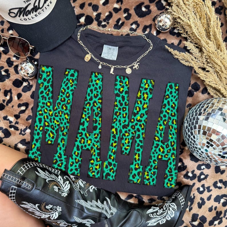 K&ouml;nnte beinhalten: Ein schwarzes T-Shirt mit dem Wort MAMA in gro&szlig;en Blockbuchstaben in einem leuchtend gr&uuml;nen und schwarzen Leopardenmuster. Das Shirt ist mit einer goldfarbenen Kette, einer schwarzen Baseballkappe und einem schwarzen Lederstiefel mit wei&szlig;er Stickerei kombiniert.