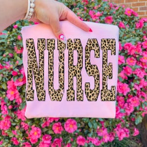 May include: A light pink sweatshirt with the word "NURSE" printed across the front in a leopard print design. The sweatshirt is held up by a hand with pink nail polish and a bracelet. The background is a bush of pink flowers.