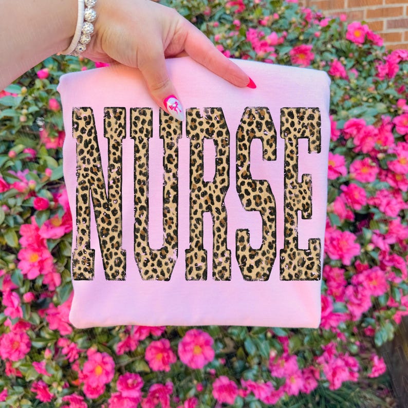 May include: A light pink sweatshirt with the word "NURSE" printed across the front in a leopard print design. The sweatshirt is held up by a hand with pink nail polish and a bracelet. The background is a bush of pink flowers.
