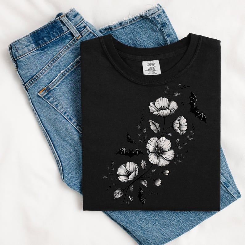 May include: Black t-shirt with a floral and bat design. The graphic features white flowers and black bats, creating a gothic aesthetic. The shirt is folded on top of a pair of blue jeans.