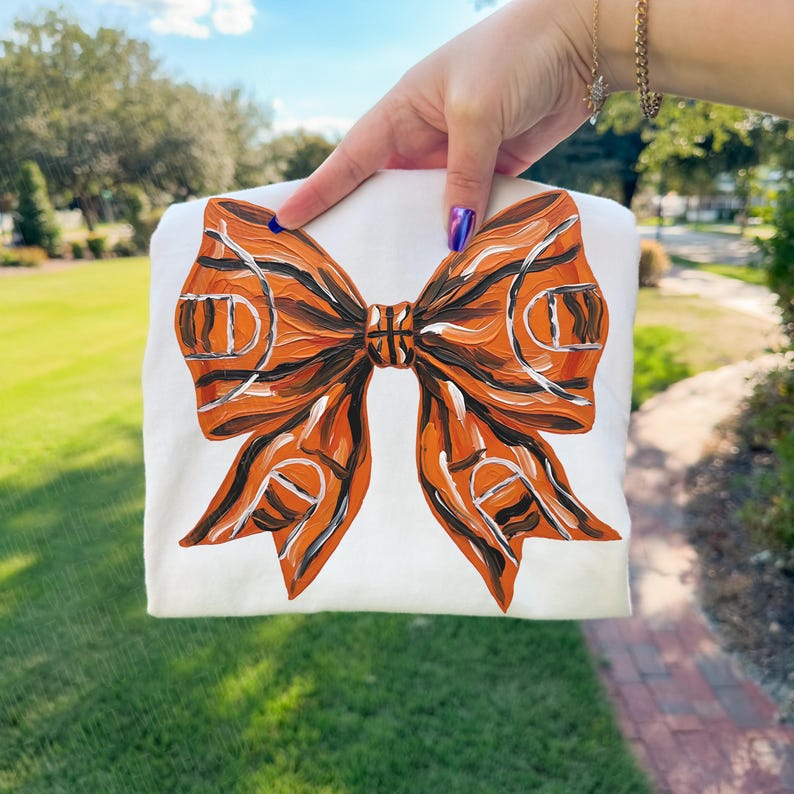 May include: White sweatshirt featuring a large, orange and black bow design. The bow has a basketball court pattern, with orange and black stripes and white lines. The sweatshirt is held up in front of a green grassy background.