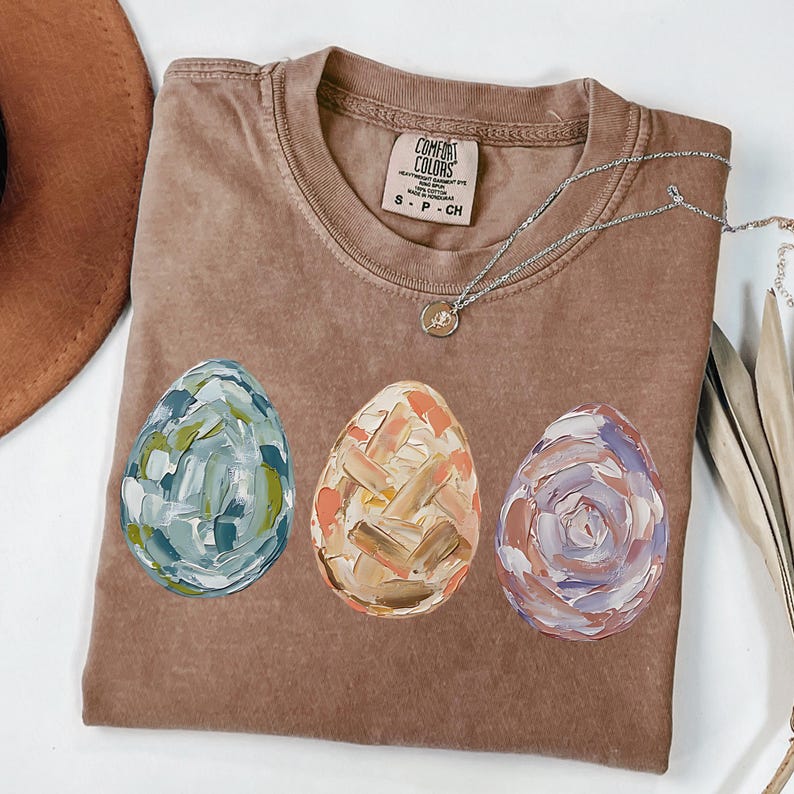 Peut inclure: T-shirt marron avec trois &oelig;ufs de P&acirc;ques peints en bleu, orange et violet. Les &oelig;ufs pr&eacute;sentent un motif textur&eacute; au pinceau. Un collier argent&eacute; avec un pendentif est pos&eacute; sur le t-shirt.