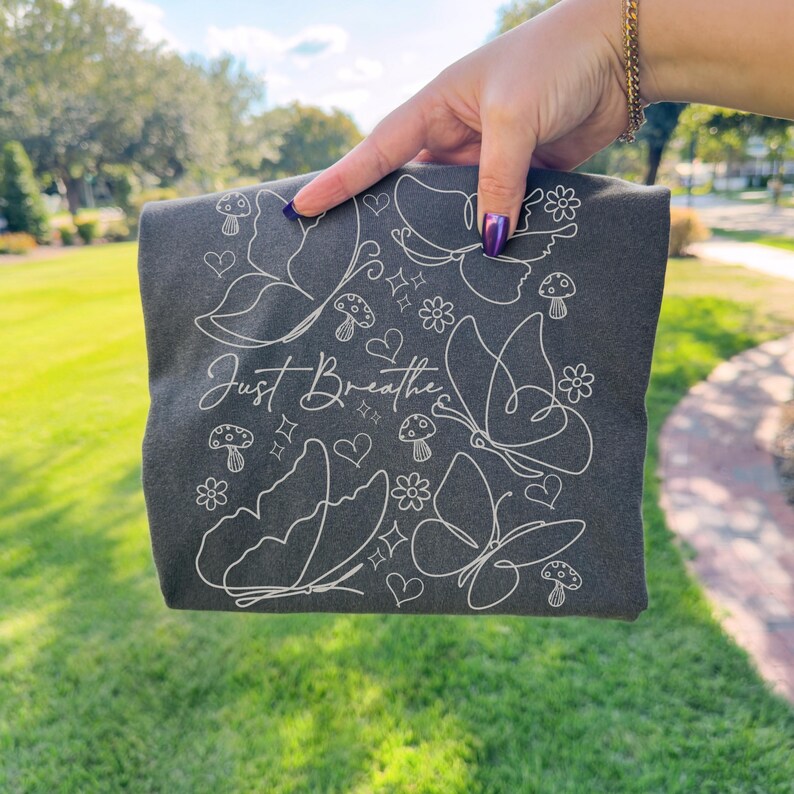 Puede incluir: Camiseta gris oscuro con un dise&ntilde;o de l&iacute;nea blanca de mariposas, setas, flores y corazones. Las palabras "Just Breathe" est&aacute;n escritas en cursiva. La camiseta se sostiene sobre un fondo de hierba verde.
