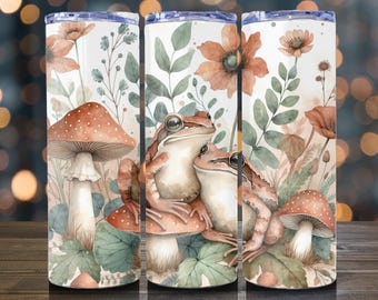 May include: A set of three stainless steel tumblers with a whimsical watercolor design. The design features two frogs sitting on a mushroom, surrounded by flowers, mushrooms, and foliage in shades of orange, green, and brown. The tumblers have a white background.