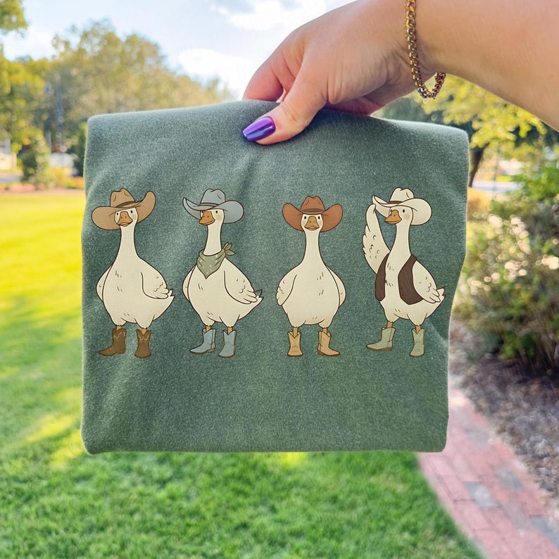 May include: Green t-shirt featuring four cartoon geese dressed as cowboys. Each goose wears a cowboy hat and boots, with one also sporting a vest and another a bandana. The geese are in various poses against a green background.