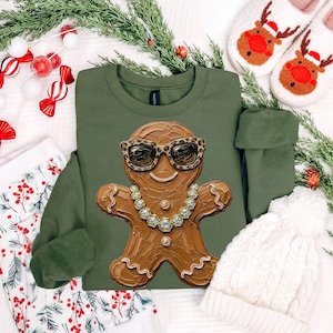 May include: An olive green crewneck sweatshirt featuring a central graphic of a brown gingerbread person wearing leopard print sunglasses and a gold chain necklace. The gingerbread figure has a textured, impasto-style paint appearance.