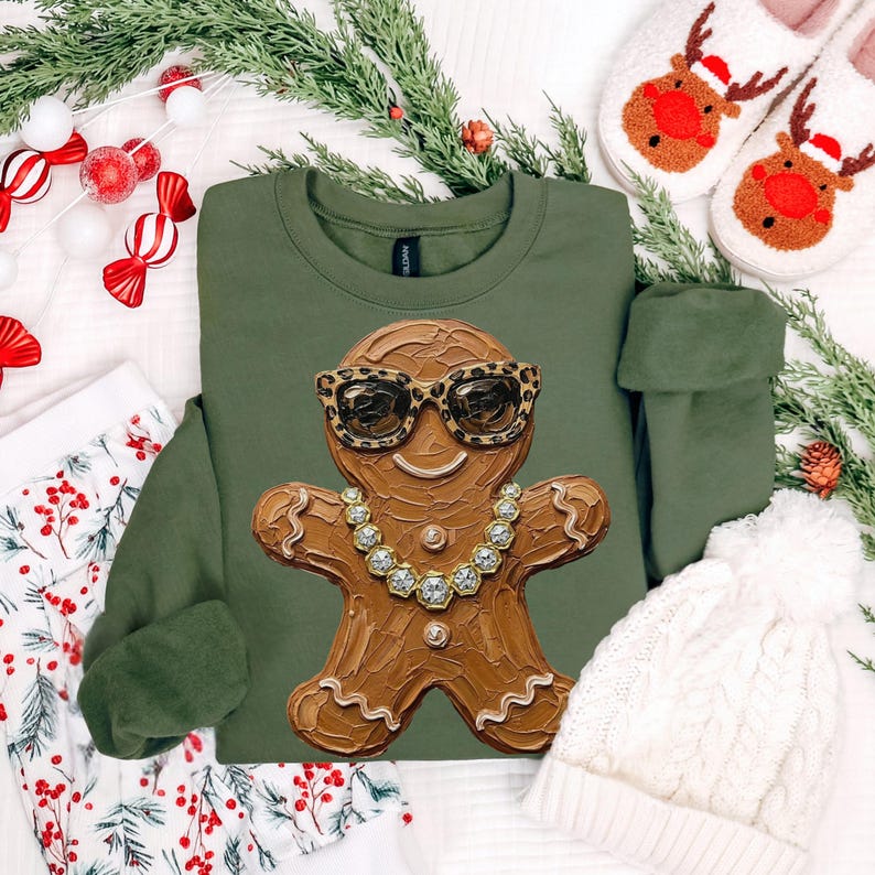 May include: An olive green crewneck sweatshirt featuring a central graphic of a brown gingerbread person wearing leopard print sunglasses and a gold chain necklace. The gingerbread figure has a textured, impasto-style paint appearance.
