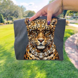 Puede incluir: Una camiseta gris oscuro con una ilustraci&oacute;n detallada de un leopardo con gafas y un collar de diamantes. El leopardo tiene un pelaje manchado en tonos marrones y dorados. La imagen se sostiene sobre un fondo de hierba verde.