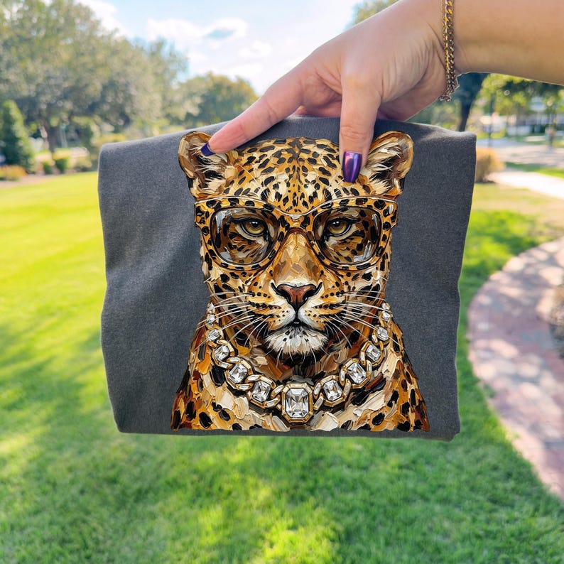 Puede incluir: Una camiseta gris oscuro con una ilustraci&oacute;n detallada de un leopardo con gafas y un collar de diamantes. El leopardo tiene un pelaje manchado en tonos marrones y dorados. La imagen se sostiene sobre un fondo de hierba verde.