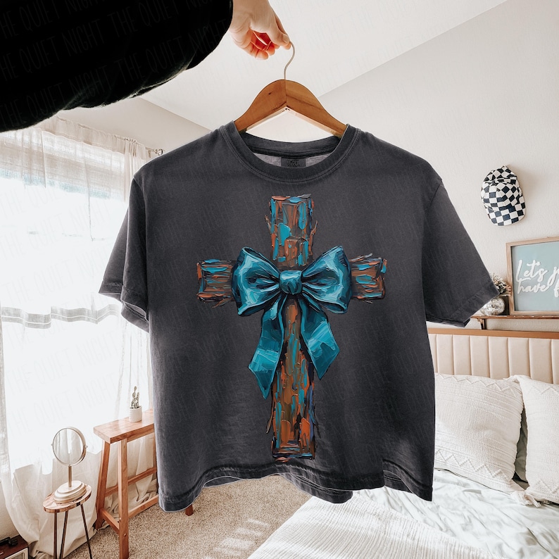 Peut inclure: T-shirt gris fonc&eacute; avec un motif de croix et un n&oelig;ud bleu. La croix a un aspect rustique et textur&eacute; avec des tons marron et orange. Le n&oelig;ud est d'un bleu vif. Le t-shirt est suspendu &agrave; un cintre en bois.