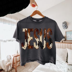K&ouml;nnte beinhalten: Dunkelgraues T-Shirt mit dem Wort "JESUS" in einem Kuhfellmuster. Das Shirt h&auml;ngt an einem Holzb&uuml;gel. Das Kuhfellmuster weist Farbt&ouml;ne von Braun, Wei&szlig; und Schwarz auf.