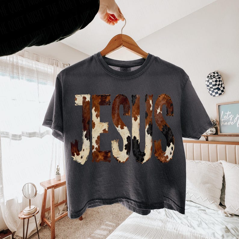 K&ouml;nnte beinhalten: Dunkelgraues T-Shirt mit dem Wort "JESUS" in einem Kuhfellmuster. Das Shirt h&auml;ngt an einem Holzb&uuml;gel. Das Kuhfellmuster weist Farbt&ouml;ne von Braun, Wei&szlig; und Schwarz auf.