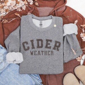 K&ouml;nnte beinhalten: Ein graues Sweatshirt mit dem Aufdruck "CIDER WEATHER" in braunen Buchstaben. Das Sweatshirt liegt auf einem rostfarbenen Kapuzenpullover und einer blauen Jeans, mit herbstlichen Dekorationen. Gem&uuml;tliches, saisonales Thema.