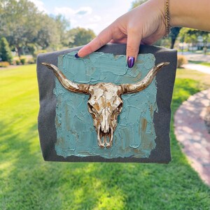 May include: A grey square textile with a turquoise painted background featuring a gold-coloured bull skull. The skull has long horns and is the focal point of the design. The artwork is held up by a hand.