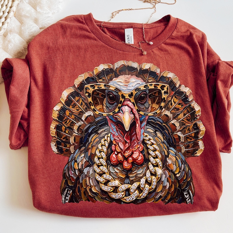 May include: A rust-colored t-shirt featuring a graphic of a turkey wearing leopard-print glasses and a thick gold chain with embedded crystals. The turkey is rendered in a textured, painterly style with brown, black, and red plumage details.