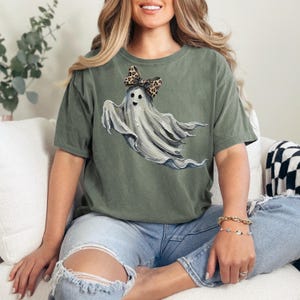 May include: An olive green t-shirt with a ghost illustration wearing a leopard print bow. The ghost is depicted with a friendly face and flowing white fabric. The shirt is a relaxed fit and is worn with blue jeans.