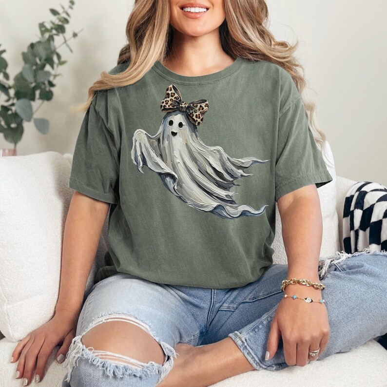 May include: An olive green t-shirt with a ghost illustration wearing a leopard print bow. The ghost is depicted with a friendly face and flowing white fabric. The shirt is a relaxed fit and is worn with blue jeans.