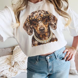 May include: A white t-shirt featuring a graphic print of a brown dapple dachshund wearing leopard print glasses and a gold-toned rhinestone collar. The dog has a mottled coat pattern with shades of brown, tan, and black, and the glasses have a distinct spotted animal print design.