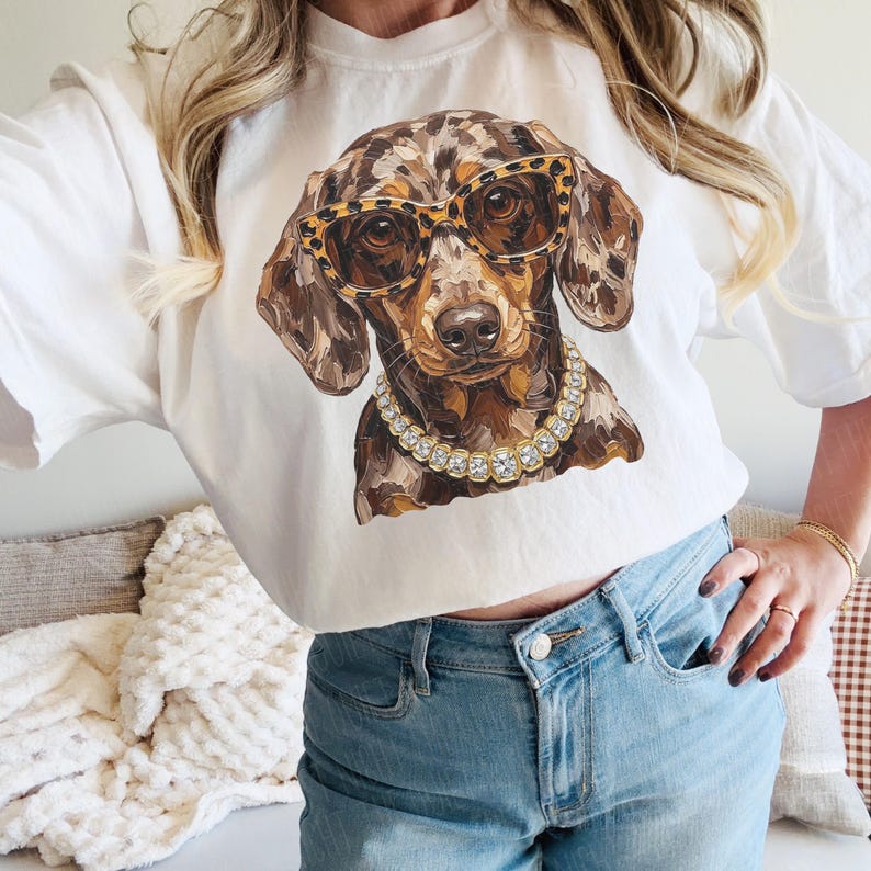 May include: A white t-shirt featuring a graphic print of a brown dapple dachshund wearing leopard print glasses and a gold-toned rhinestone collar. The dog has a mottled coat pattern with shades of brown, tan, and black, and the glasses have a distinct spotted animal print design.