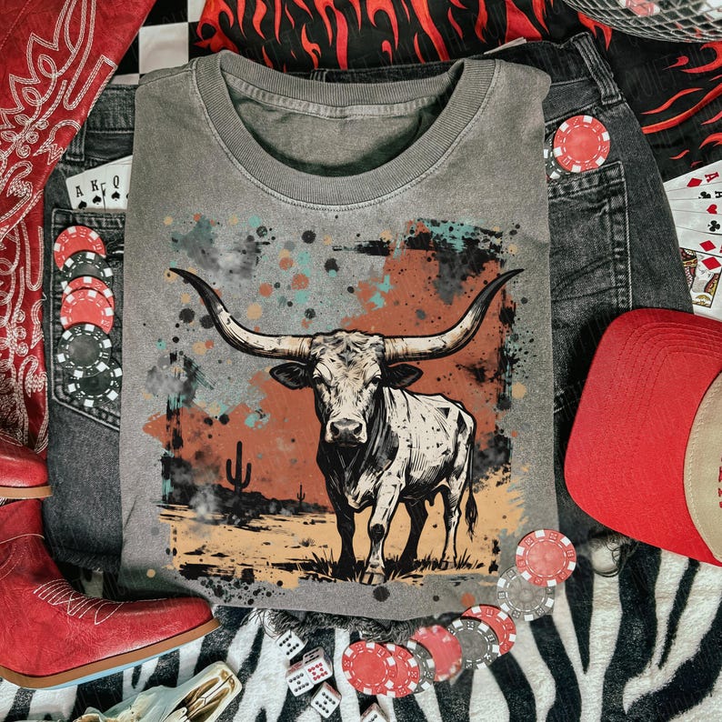 May include: Gray crew neck t-shirt featuring a longhorn steer illustration with a desert landscape background. The image is surrounded by denim jeans, a red cap, red cowboy boots, poker chips, dice, and playing cards.