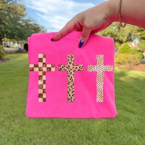 May include: A bright pink t-shirt featuring three cross designs. The crosses have unique patterns: a checkered design, a leopard print, and a dotted pattern. The shirt is held up against a green grassy background.