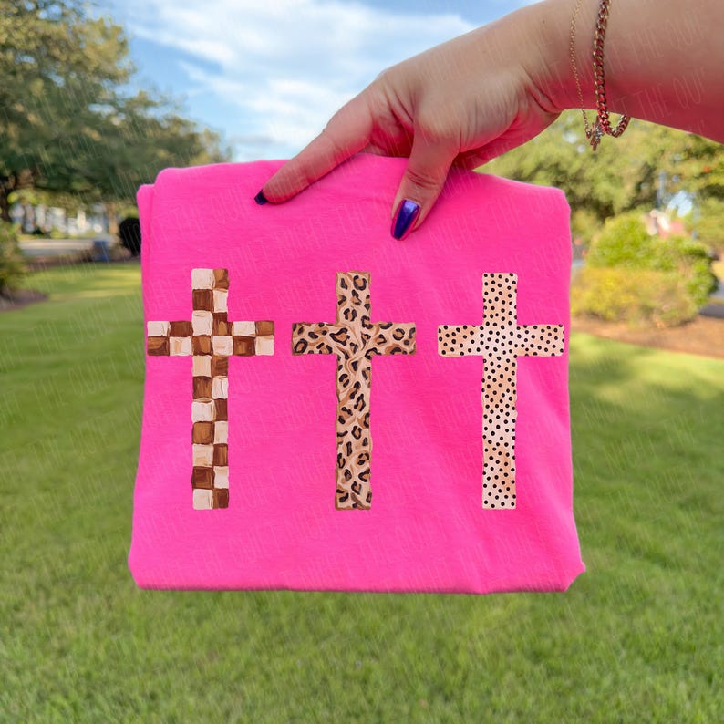 May include: A bright pink t-shirt featuring three cross designs. The crosses have unique patterns: a checkered design, a leopard print, and a dotted pattern. The shirt is held up against a green grassy background.