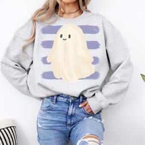 May include: A light gray crewneck sweatshirt featuring a central graphic of a cream-colored ghost with a simple smiling face, positioned over four horizontal, distressed-texture purple brushstroke stripes across the chest area.