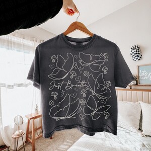 Puede incluir: Camiseta gris oscuro con un dise&ntilde;o de mariposas y flores blancas. Las palabras "Just Breathe" est&aacute;n escritas en cursiva. La camiseta est&aacute; colgada en una percha de madera.