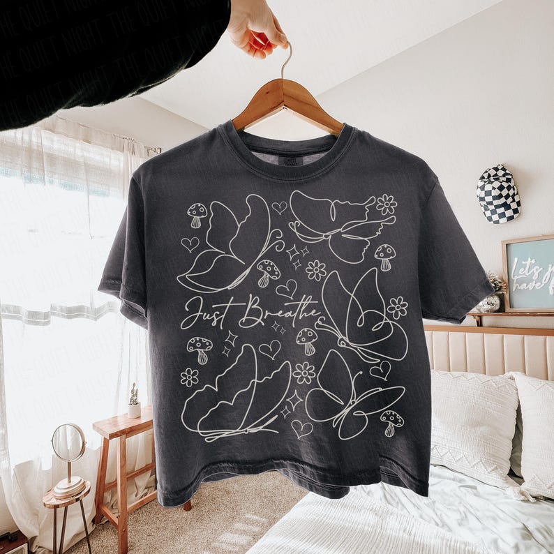 Puede incluir: Camiseta gris oscuro con un dise&ntilde;o de mariposas y flores blancas. Las palabras "Just Breathe" est&aacute;n escritas en cursiva. La camiseta est&aacute; colgada en una percha de madera.
