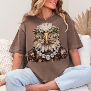 May include: Brown t-shirt featuring a detailed illustration of an eagle wearing leopard print glasses and a diamond necklace. The eagle's feathers are rendered in shades of brown, beige, and white. The shirt is a relaxed fit.