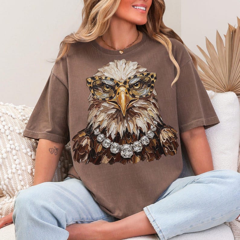 May include: Brown t-shirt featuring a detailed illustration of an eagle wearing leopard print glasses and a diamond necklace. The eagle's feathers are rendered in shades of brown, beige, and white. The shirt is a relaxed fit.