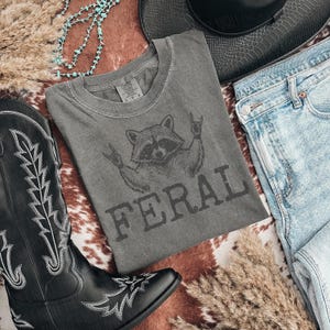 May include: A gray t-shirt with a raccoon graphic and the word "FERAL" in dark gray lettering. The shirt is laid out with a pair of light blue jeans, a black cowboy boot, and a black hat. A turquoise necklace and dried grass are also in the image.