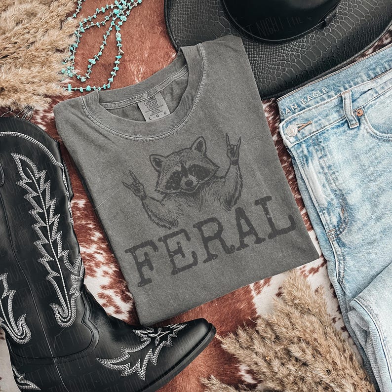 May include: A gray t-shirt with a raccoon graphic and the word "FERAL" in dark gray lettering. The shirt is laid out with a pair of light blue jeans, a black cowboy boot, and a black hat. A turquoise necklace and dried grass are also in the image.