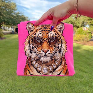 K&ouml;nnte beinhalten: Ein leuchtend pinkfarbenes T-Shirt mit einer Illustration eines Tigers, der eine Sonnenbrille und eine Diamantkette tr&auml;gt. Der Tiger hat orange und schwarze Streifen. Der Hintergrund ist leuchtend pink.