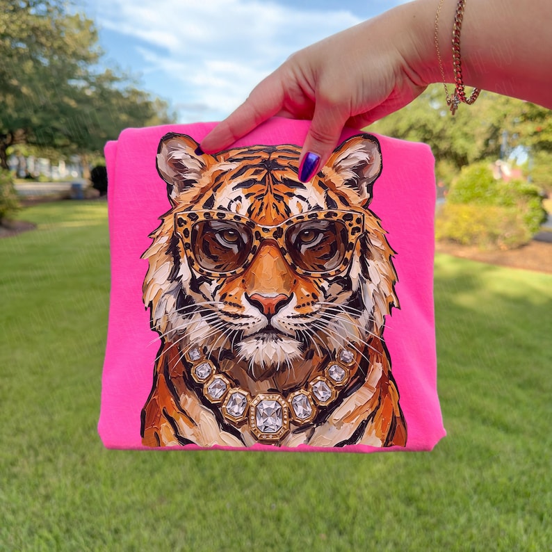K&ouml;nnte beinhalten: Ein leuchtend pinkfarbenes T-Shirt mit einer Illustration eines Tigers, der eine Sonnenbrille und eine Diamantkette tr&auml;gt. Der Tiger hat orange und schwarze Streifen. Der Hintergrund ist leuchtend pink.