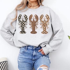 May include: A light gray sweatshirt featuring three lobster designs. Each lobster has a different pattern: black and white checkered, leopard print, and tan with black polka dots. The sweatshirt is paired with blue jeans.