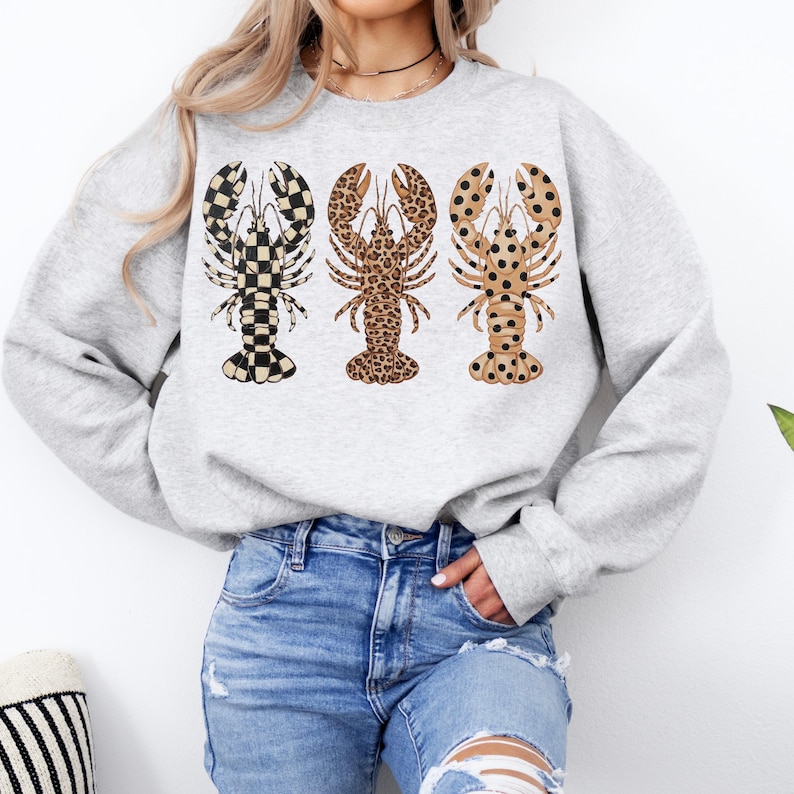 May include: A light gray sweatshirt featuring three lobster designs. Each lobster has a different pattern: black and white checkered, leopard print, and tan with black polka dots. The sweatshirt is paired with blue jeans.