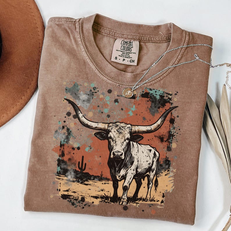 May include: A brown Comfort Colors t-shirt featuring a graphic of a longhorn steer with large horns. The steer is set against a desert landscape with a cactus and colorful paint splatters. A silver necklace with a pendant is draped on the shirt.