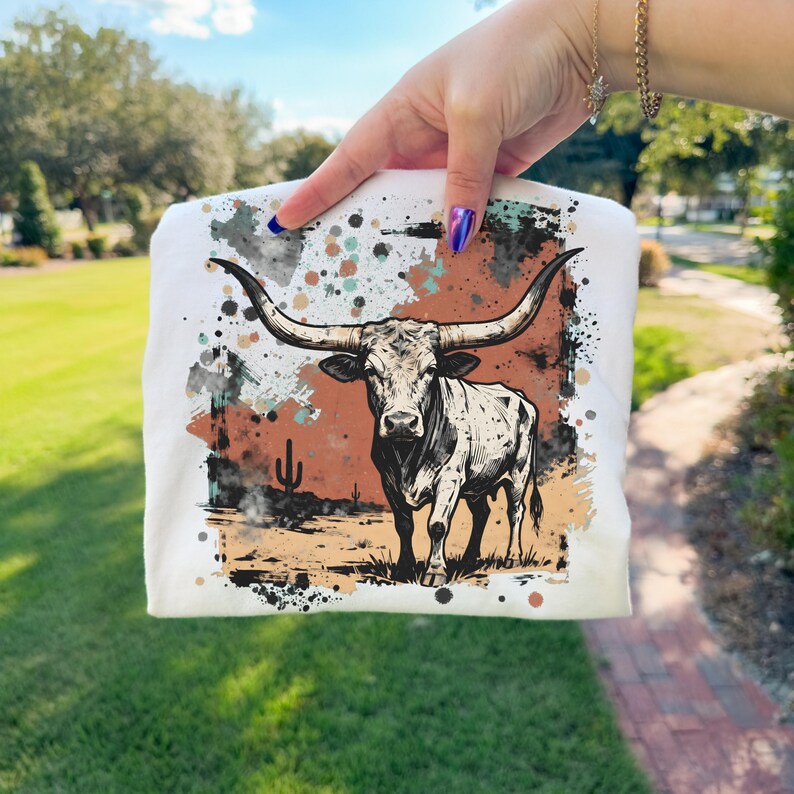 May include: White t-shirt featuring a detailed illustration of a longhorn steer with large horns, set against a desert landscape with a cactus. The design includes brown, orange, and teal paint splatters.