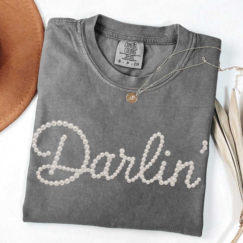 Pu&ograve; includere: Una maglietta grigia Comfort Colors con la parola "Darlin'" scritta con perle bianche. Sono visibili anche una collana d'oro con un ciondolo e un cappello marrone.