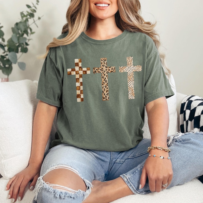 May include: Olive green t-shirt featuring three crosses with different patterns: checkered, leopard print, and dotted. The crosses are in shades of brown and tan. The shirt is a casual, comfortable style.