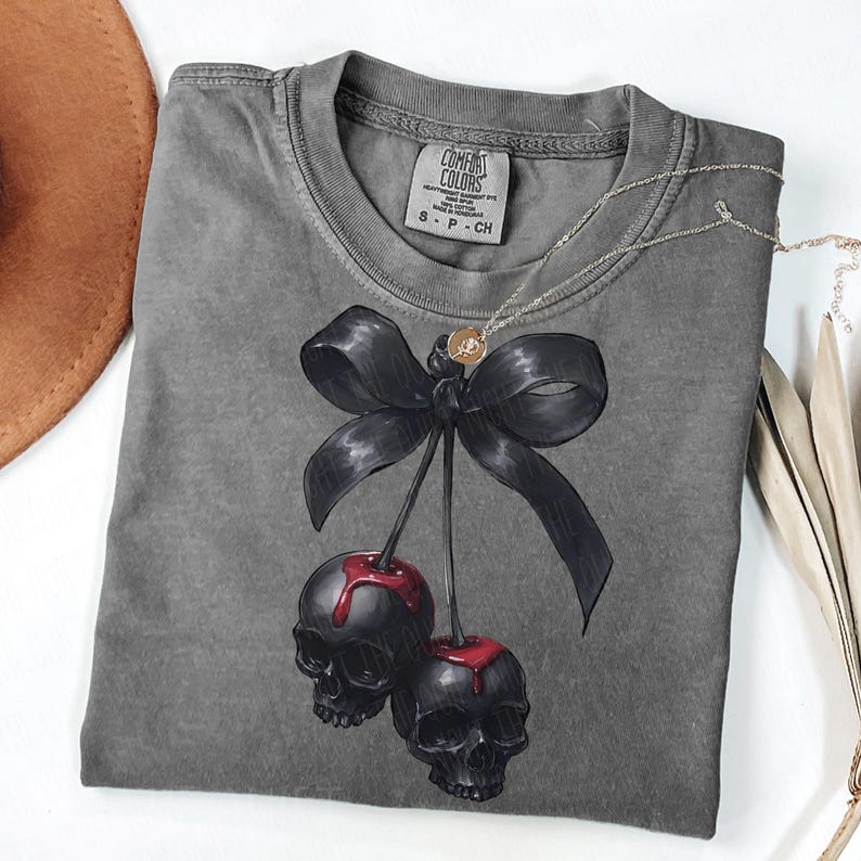 May include: A grey Comfort Colors t-shirt with a black bow and two cherries with skull faces and red juice. A silver necklace with a small pendant is draped across the shirt. The shirt has a small tag that reads "S P CH".