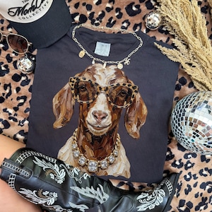 May include: A dark gray t-shirt featuring a painted illustration of a goat wearing leopard print sunglasses and a chunky gold rhinestone necklace. The goat has brown and white fur. A gold chain necklace rests on the collar of the shirt.