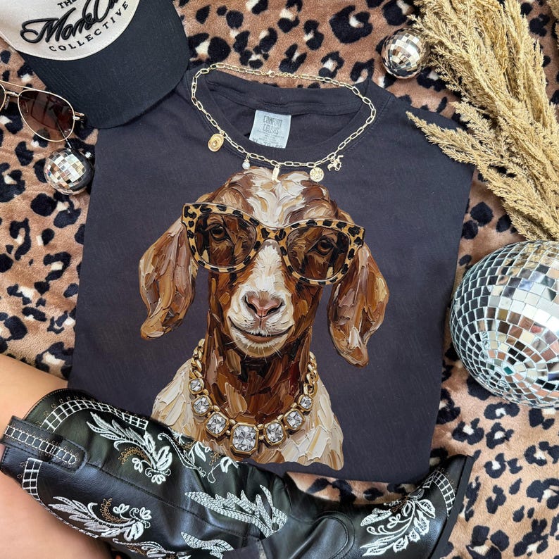 May include: A dark gray t-shirt featuring a painted illustration of a goat wearing leopard print sunglasses and a chunky gold rhinestone necklace. The goat has brown and white fur. A gold chain necklace rests on the collar of the shirt.