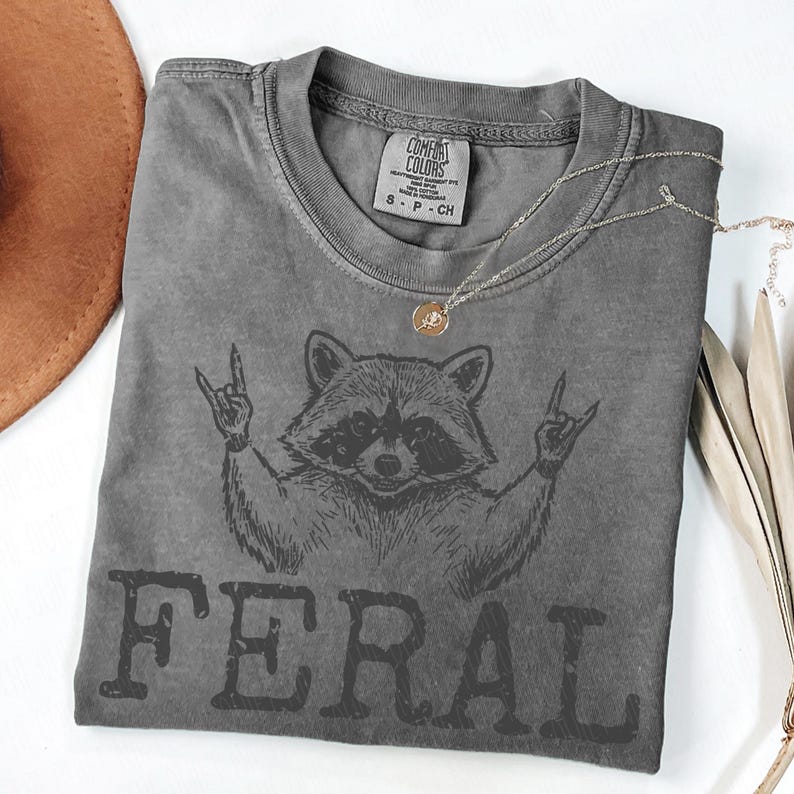 May include: A gray t-shirt featuring a raccoon graphic with the word "FERAL" printed below. The raccoon is making a rock and roll hand gesture. A gold necklace is draped across the shirt.
