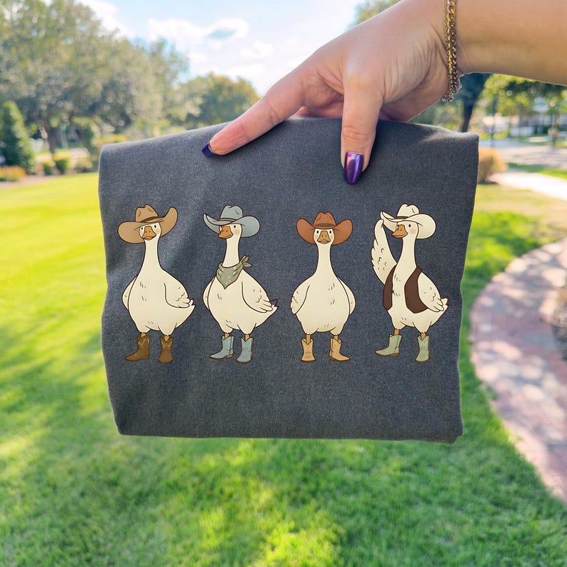 May include: Dark gray t-shirt featuring four cartoon geese dressed as cowboys. Each goose wears a cowboy hat, boots, and some with bandanas or vests. The geese are in various poses, adding a playful touch to the design.