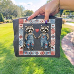 Pu&ograve; includere: T-shirt grigio scuro con un'illustrazione ad acquerello di due procioni che si tengono per mano e un palloncino a forma di cuore. I procioni indossano salopette blu con dettagli a cuore. Il disegno &egrave; incorniciato da un bordo patchwork di cuori e forme geometriche.