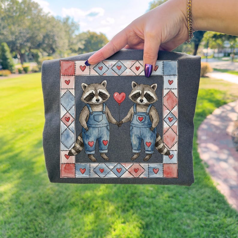 Pu&ograve; includere: T-shirt grigio scuro con un'illustrazione ad acquerello di due procioni che si tengono per mano e un palloncino a forma di cuore. I procioni indossano salopette blu con dettagli a cuore. Il disegno &egrave; incorniciato da un bordo patchwork di cuori e forme geometriche.