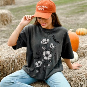 May include: Dark gray t-shirt featuring a floral and bat design. The shirt has a vintage wash look. The person is wearing a corduroy orange cap with the words "PUMPKIN SPICE" embroidered on the front.