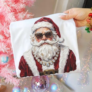 May include: A white t-shirt featuring a painterly graphic of Santa Claus wearing leopard print sunglasses and a chunky crystal-style necklace. The artwork displays thick, textured brushstrokes in shades of red, white, and brown.